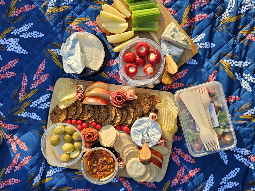 A charcuterie board with a variety of crackers, vegetables, and cured meats sits on top of a dark blue floral quilted picnic blanket. It's well lit and sunny.