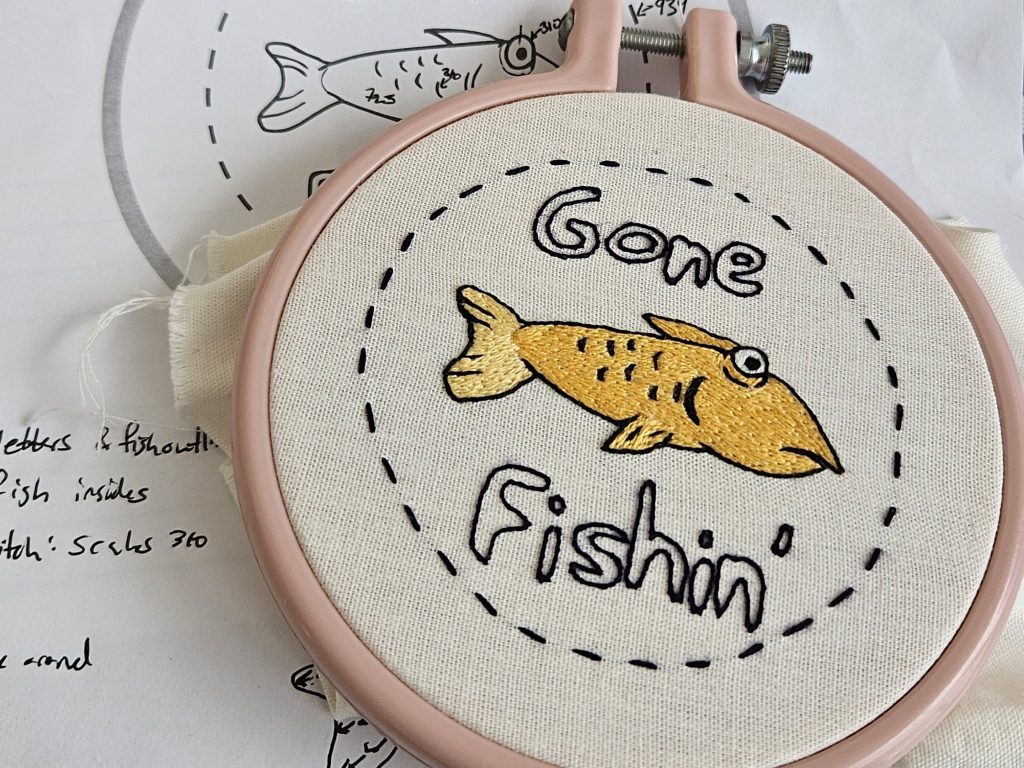 A hand embroidered fish from the Club Penguin fishing minigame sits in a pink embroidery hoop. Behind it is its embroidery pattern with notes written on it.