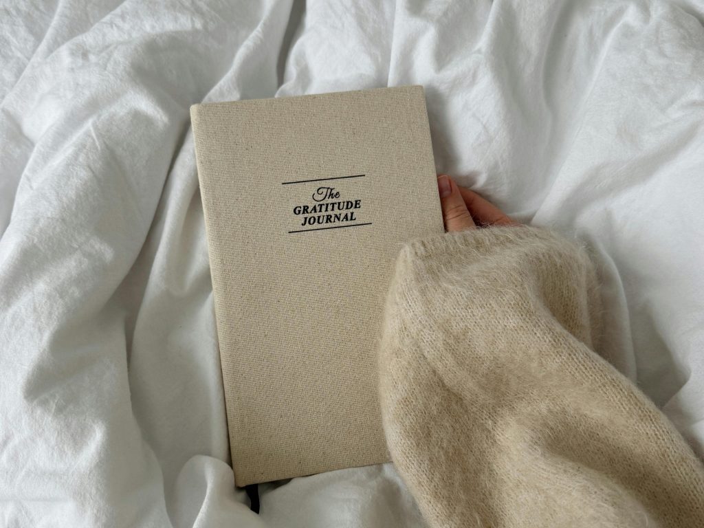 a woman's hand holds a gratitude journal atop a a cozy white duvet. The journal is beige and the hand is wearing a fuzzy beige sweater. 