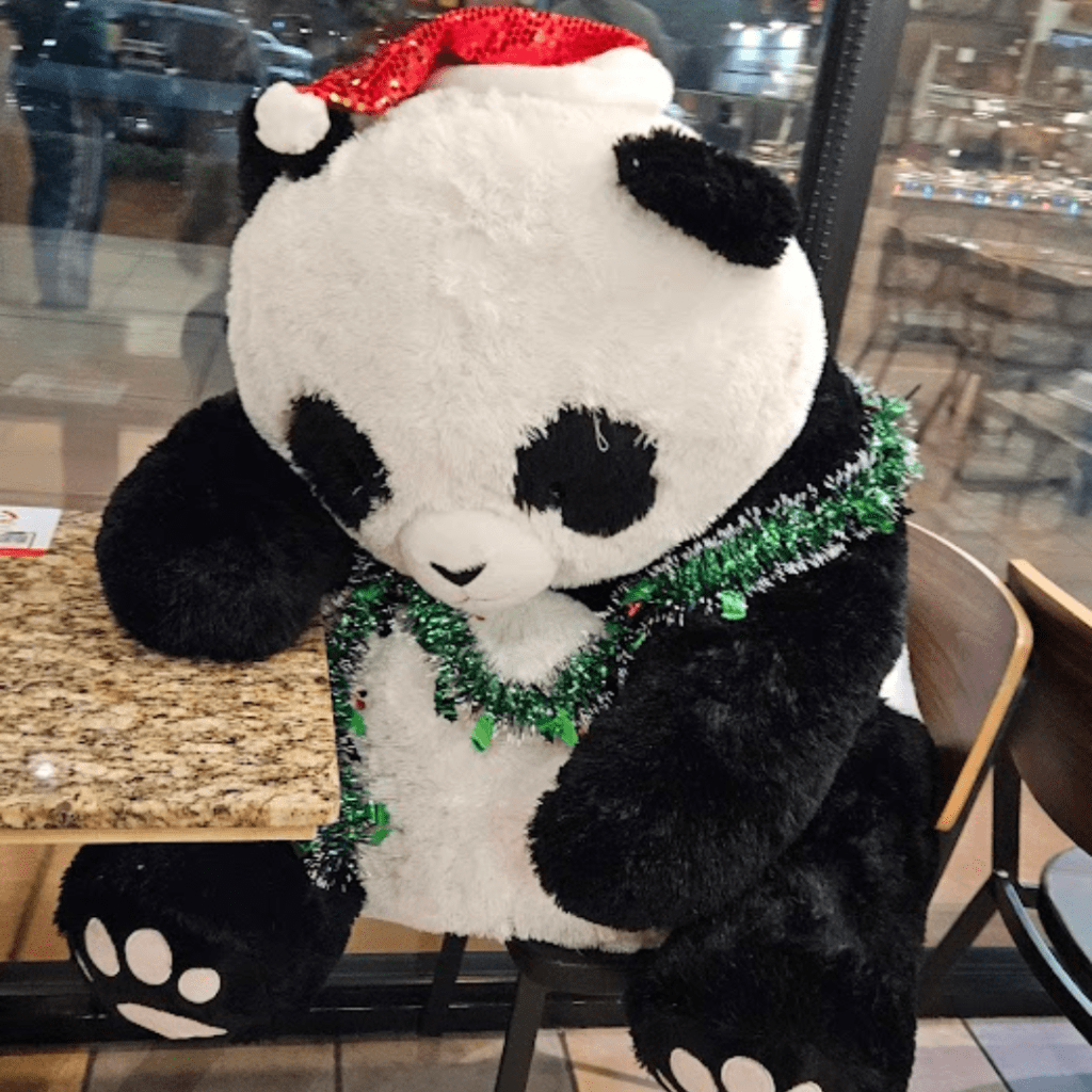 a stuffed panda looking totally exhausted and done with life. it's wearing a santa hat and a festive garland around its torso.